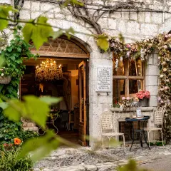 Urokliwa kamienna fasada w Saint-Paul-de-Vence, ozdobiona kwiatami i pnączami. W otwartych drzwiach wisi kryształowy żyrandol.