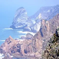 Skaliste wybrzeże Cabo da Roca, z falami rozbijającymi się o klify, w oddali widać mgliste góry.