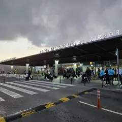 Ludzie z bagażami wchodzą do portu lotniczego Pafos pod neonowym napisem.
