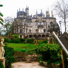 Kamienna rzeźba w ogrodzie Quinta da Regaleira, z widokiem na ozdobny pałac i schody prowadzące do niego.