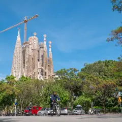 Majestatyczna La Sagrada Familia w Barcelonie, wciąż w budowie, góruje nad zielonym parkiem i ulicą, gdzie rowerzysta przemierza drogę.