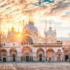 Wschód słońca nad Bazyliką św. Marka na Piazza San Marco. Złote promienie przebijają się przez chmury, oświetlając wenecką architekturę.
