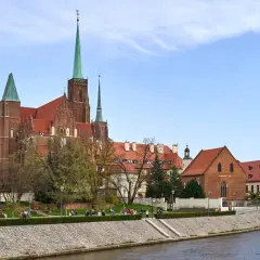 Ostrów Tumski we Wrocławiu z katedrą i zielonymi wieżami, nad rzeką, z ludźmi spacerującymi po bulwarze.