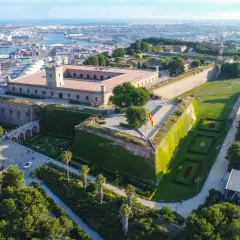 Zamek Castell de Montjuïc góruje nad portem w Barcelonie. Otoczony zielenią, z flagą Katalonii powiewającą na wietrze.