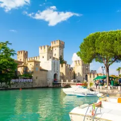 Malowniczy widok na Sirmione Castello Scaligero nad turkusową wodą, z łodziami zacumowanymi przy brzegu i zielonymi drzewami.