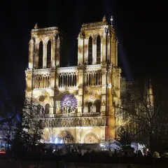 Katedra Notre Dame w Paryżu nocą, oświetlona reflektorami. Widać tłum ludzi oglądających spektakl.