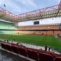 Pusta trybuna stadionu San Siro z zieloną murawą i pomarańczowymi oraz niebieskimi siedzeniami.