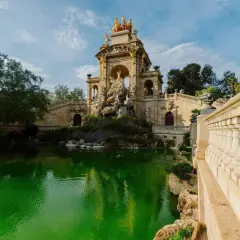 Monumentalna fontanna z rydwanem na szczycie, otoczona zielonym stawem i kamiennymi schodami w Parc de la Ciutadella.
