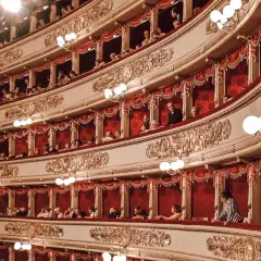 Wnętrze La Scala w Mediolanie, z widownią w lożach, ozdobione złotymi zdobieniami i czerwonymi zasłonami.