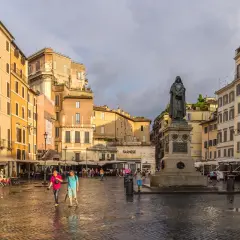 Pomnik na placu Campo de' Fiori w Rzymie, otoczony kolorowymi budynkami i ludźmi spacerującymi po mokrym bruku.