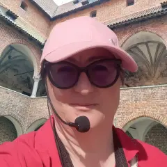 Kobieta w czapce i okularach przeciwsłonecznych zwiedza z przewodnikiem z mikrofonem zamek w Lidzbarku Warmińskim, odkrywając jego atrakcje.