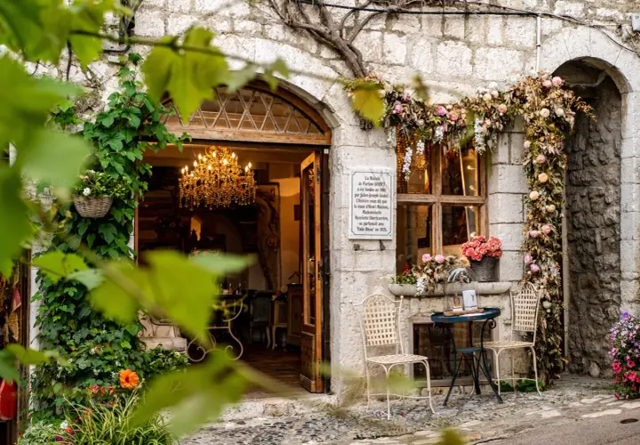 Urokliwa kamienna fasada w Saint-Paul-de-Vence, ozdobiona kwiatami i pnączami. W otwartych drzwiach wisi kryształowy żyrandol.