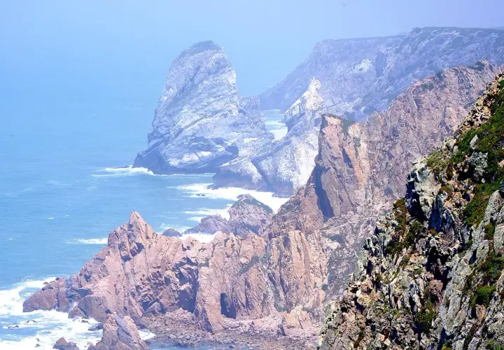 Skaliste wybrzeże Cabo da Roca, z falami rozbijającymi się o klify, w oddali widać mgliste góry.