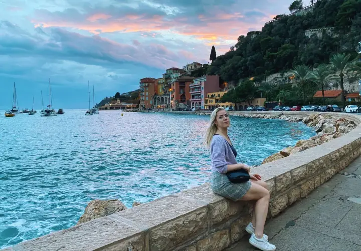 Kobieta siedzi na kamiennym murku nad morzem w Villefranche-sur-Mer, podziwiając zachód słońca i żaglówki.