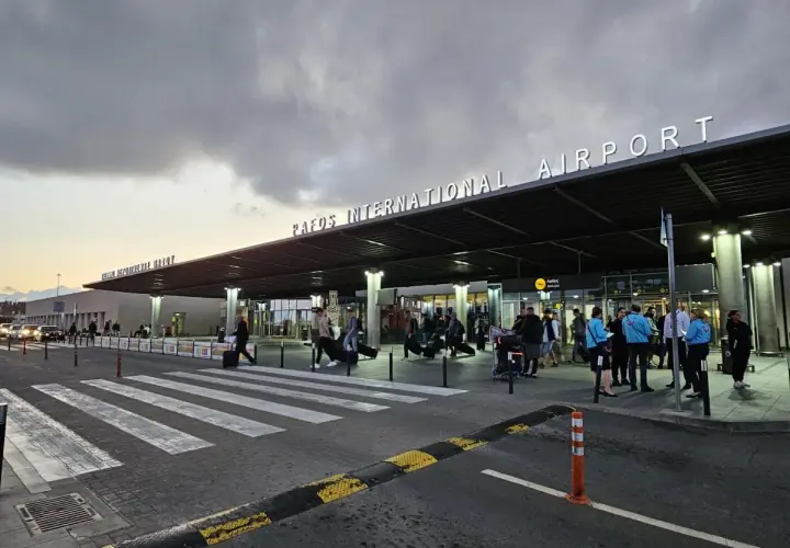Ludzie z bagażami wchodzą do portu lotniczego Pafos pod neonowym napisem.