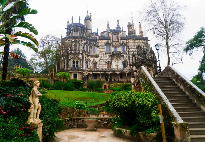 Kamienna rzeźba w ogrodzie Quinta da Regaleira, z widokiem na ozdobny pałac i schody prowadzące do niego.