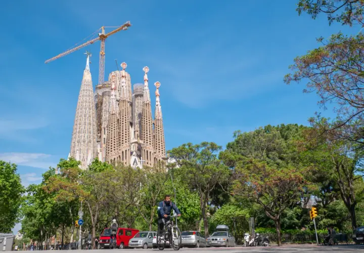 Majestatyczna La Sagrada Familia w Barcelonie, wciąż w budowie, góruje nad zielonym parkiem i ulicą, gdzie rowerzysta przemierza drogę.