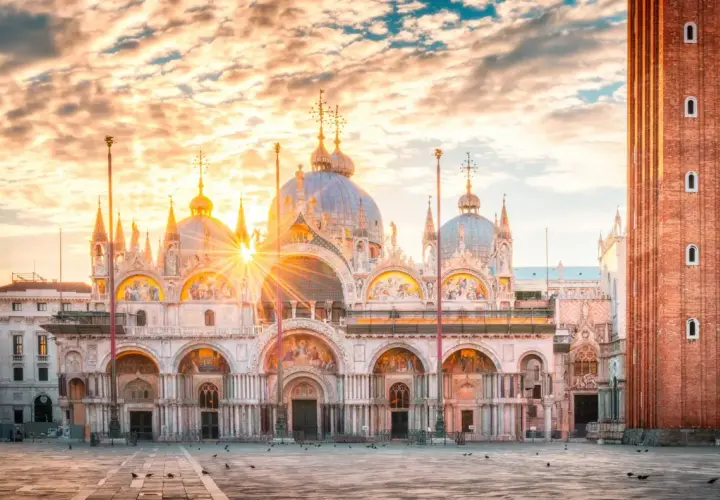 Wschód słońca nad Bazyliką św. Marka na Piazza San Marco. Złote promienie przebijają się przez chmury, oświetlając wenecką architekturę.