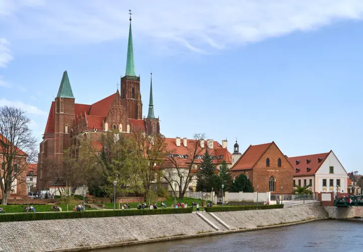 Ostrów Tumski we Wrocławiu z katedrą i zielonymi wieżami, nad rzeką, z ludźmi spacerującymi po bulwarze.