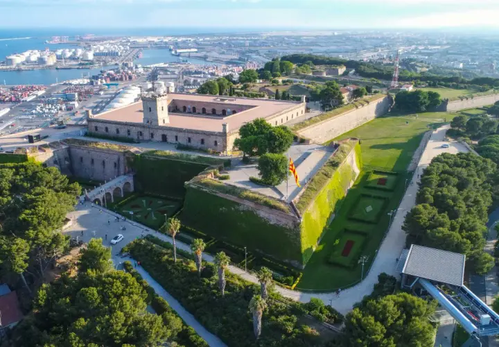 Zamek Castell de Montjuïc góruje nad portem w Barcelonie. Otoczony zielenią, z flagą Katalonii powiewającą na wietrze.