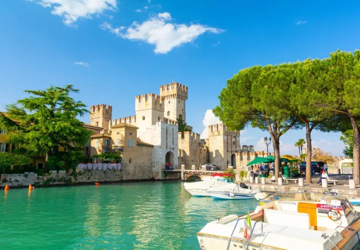 Malowniczy widok na Sirmione Castello Scaligero nad turkusową wodą, z łodziami zacumowanymi przy brzegu i zielonymi drzewami.