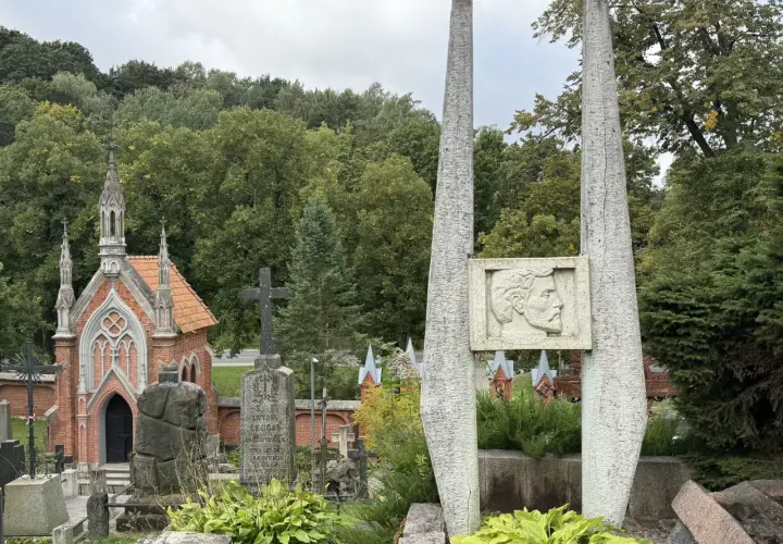 Kamienne obeliski i neogotycka kaplica na cmentarzu na Rossie. W tle zieleń drzew i pochmurne niebo.