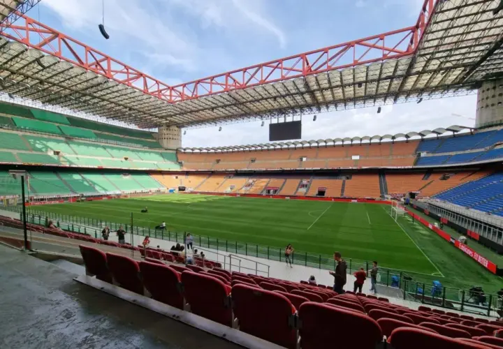 Pusta trybuna stadionu San Siro z zieloną murawą i pomarańczowymi oraz niebieskimi siedzeniami.