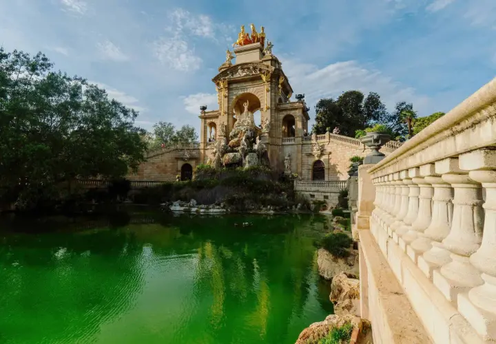 Monumentalna fontanna z rydwanem na szczycie, otoczona zielonym stawem i kamiennymi schodami w Parc de la Ciutadella.