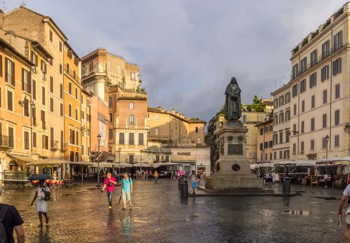 Pomnik na placu Campo de' Fiori w Rzymie, otoczony kolorowymi budynkami i ludźmi spacerującymi po mokrym bruku.