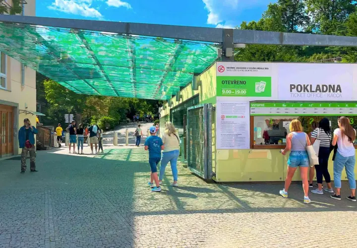 Wejście do ogrodu zoologicznego w Pradze. Ludzie stoją w kolejce do kasy biletowej pod zielonym zadaszeniem.