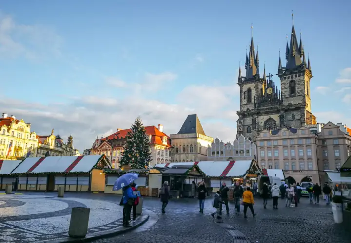 Rynek Staromiejski w Pradze, z choinką i straganami świątecznymi, podziwiany przez spacerujących ludzi.