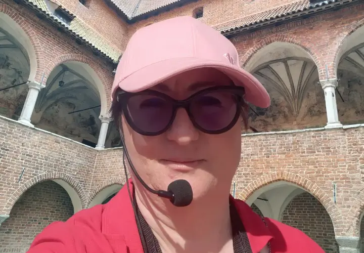 Kobieta w czapce i okularach przeciwsłonecznych zwiedza z przewodnikiem z mikrofonem zamek w Lidzbarku Warmińskim, odkrywając jego atrakcje.