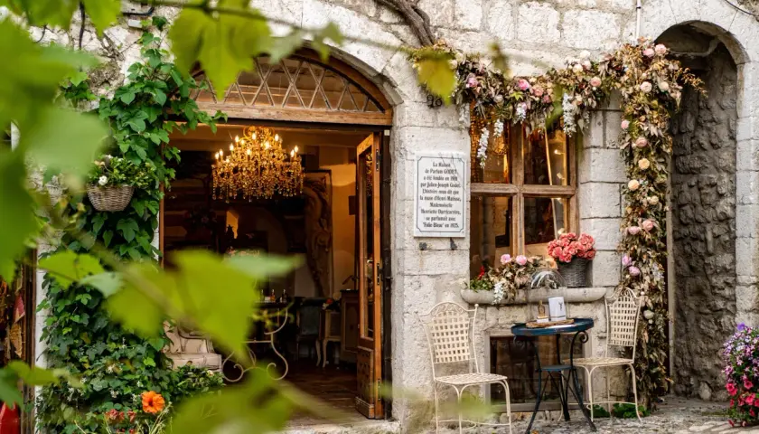 Urokliwa kamienna fasada w Saint-Paul-de-Vence, ozdobiona kwiatami i pnączami. W otwartych drzwiach wisi kryształowy żyrandol.