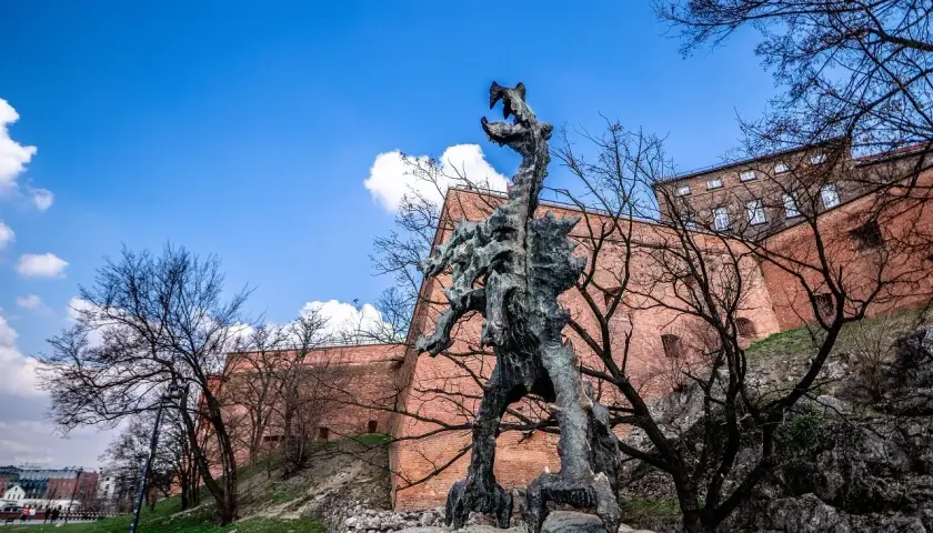 Smok Wawelski Kraków, groźny posąg smoka u stóp Wawelu, na tle ceglanych murów i błękitnego nieba.