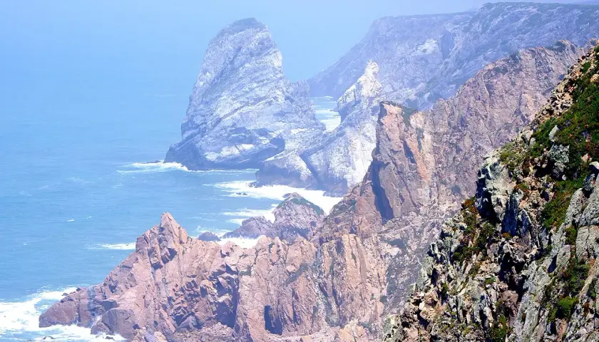 Skaliste wybrzeże Cabo da Roca, z falami rozbijającymi się o klify, w oddali widać mgliste góry.