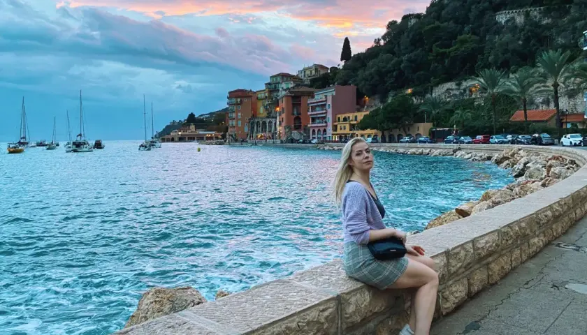 Kobieta siedzi na kamiennym murku nad morzem w Villefranche-sur-Mer, podziwiając zachód słońca i żaglówki.