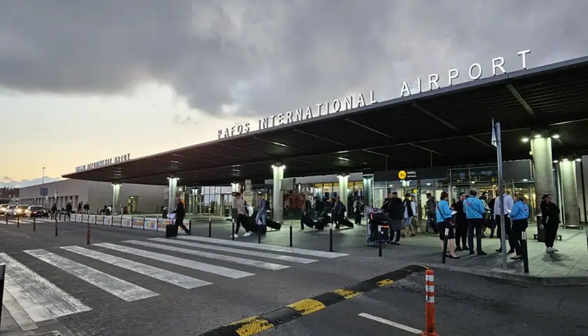 Ludzie z bagażami wchodzą do portu lotniczego Pafos pod neonowym napisem.