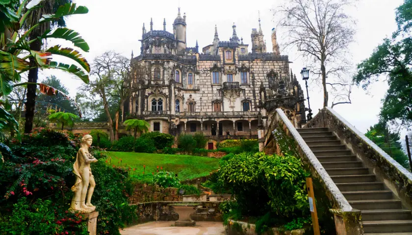 Kamienna rzeźba w ogrodzie Quinta da Regaleira, z widokiem na ozdobny pałac i schody prowadzące do niego.