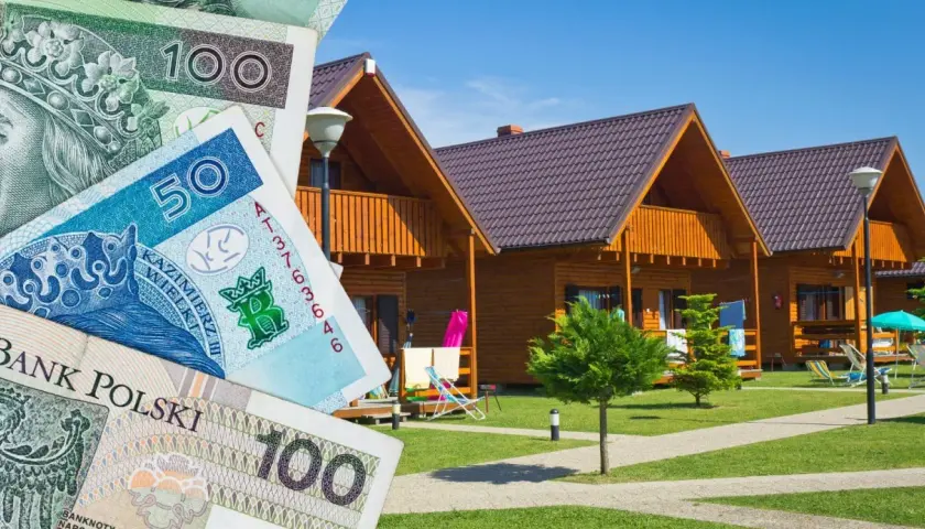 Polskie banknoty przed drewnianymi domkami letniskowymi. Zastanawiasz się, gdzie najlepiej rezerwować noclegi, by wakacje były udane i nie zrujnowały budżetu?