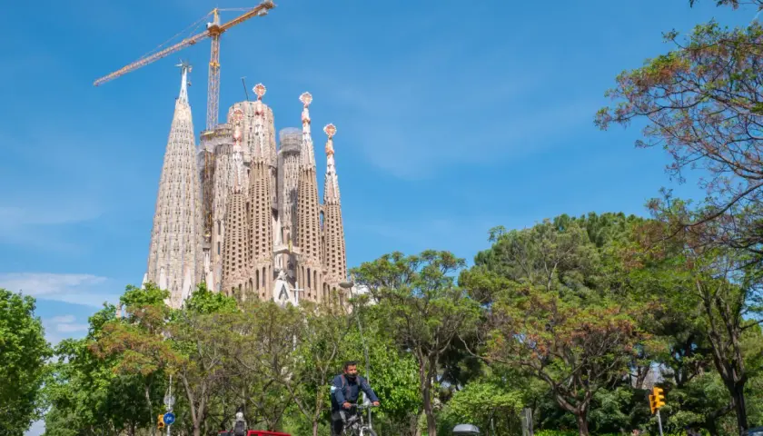 Majestatyczna La Sagrada Familia w Barcelonie, wciąż w budowie, góruje nad zielonym parkiem i ulicą, gdzie rowerzysta przemierza drogę.