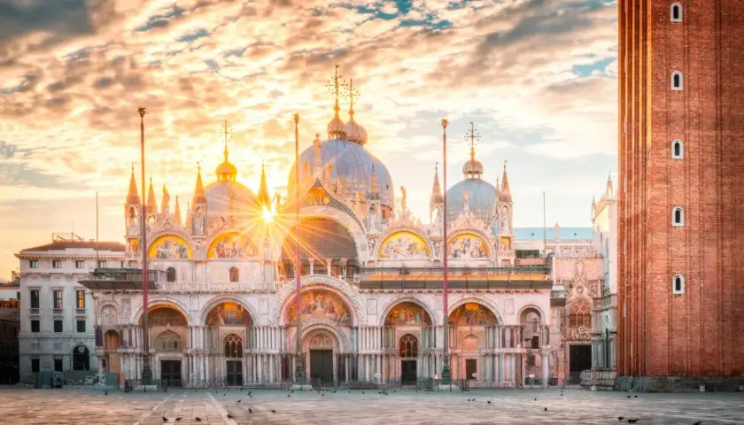 Wschód słońca nad Bazyliką św. Marka na Piazza San Marco. Złote promienie przebijają się przez chmury, oświetlając wenecką architekturę.