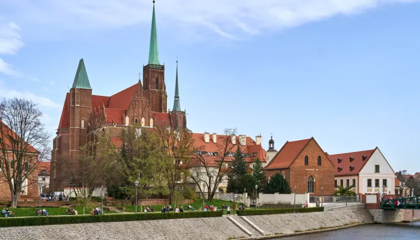 Ostrów Tumski we Wrocławiu z katedrą i zielonymi wieżami, nad rzeką, z ludźmi spacerującymi po bulwarze.
