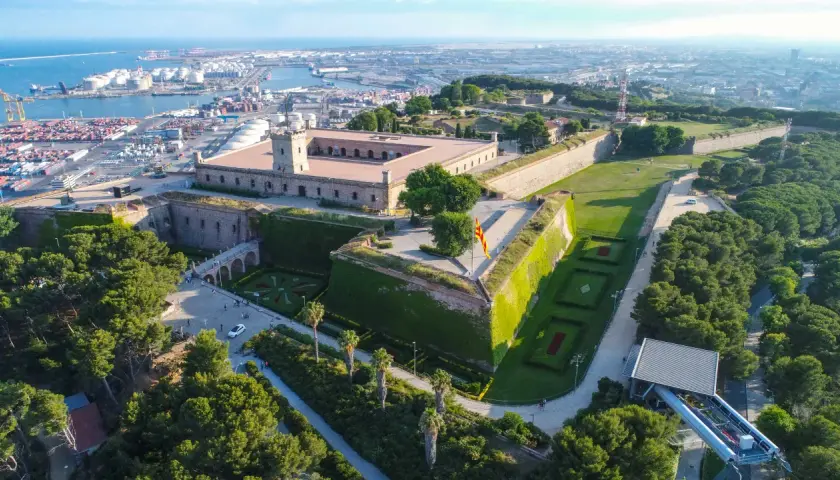 Zamek Castell de Montjuïc góruje nad portem w Barcelonie. Otoczony zielenią, z flagą Katalonii powiewającą na wietrze.