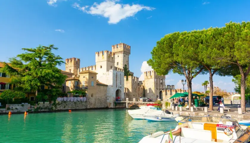 Malowniczy widok na Sirmione Castello Scaligero nad turkusową wodą, z łodziami zacumowanymi przy brzegu i zielonymi drzewami.