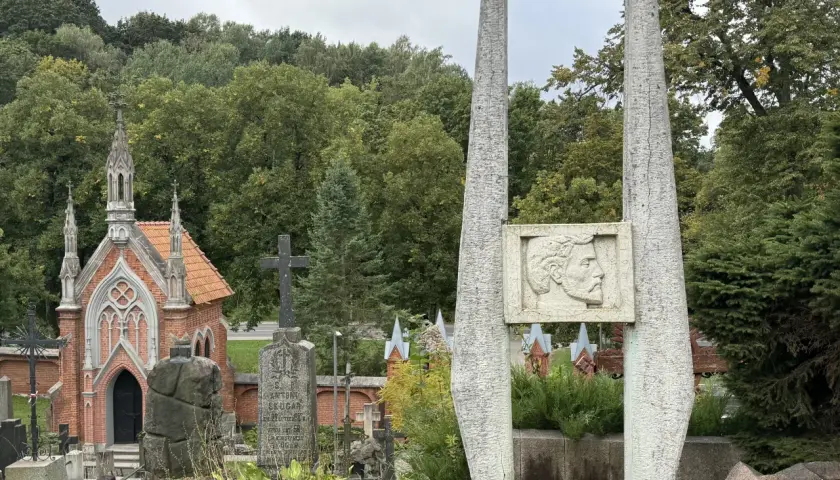 Kamienne obeliski i neogotycka kaplica na cmentarzu na Rossie. W tle zieleń drzew i pochmurne niebo.