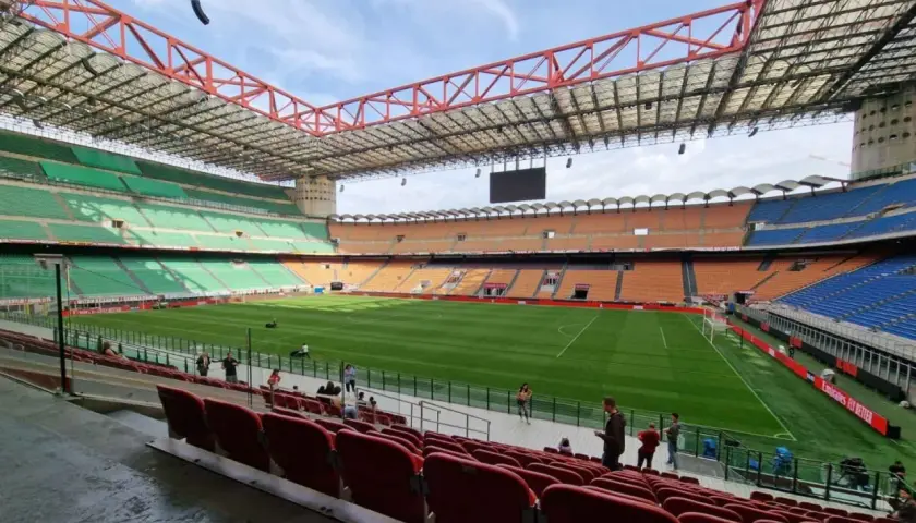 Pusta trybuna stadionu San Siro z zieloną murawą i pomarańczowymi oraz niebieskimi siedzeniami.
