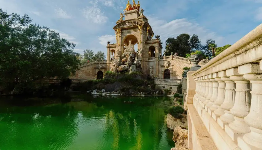 Monumentalna fontanna z rydwanem na szczycie, otoczona zielonym stawem i kamiennymi schodami w Parc de la Ciutadella.