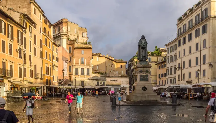 Pomnik na placu Campo de' Fiori w Rzymie, otoczony kolorowymi budynkami i ludźmi spacerującymi po mokrym bruku.