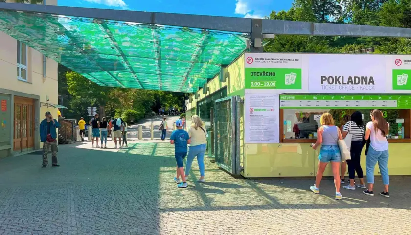 Wejście do ogrodu zoologicznego w Pradze. Ludzie stoją w kolejce do kasy biletowej pod zielonym zadaszeniem.