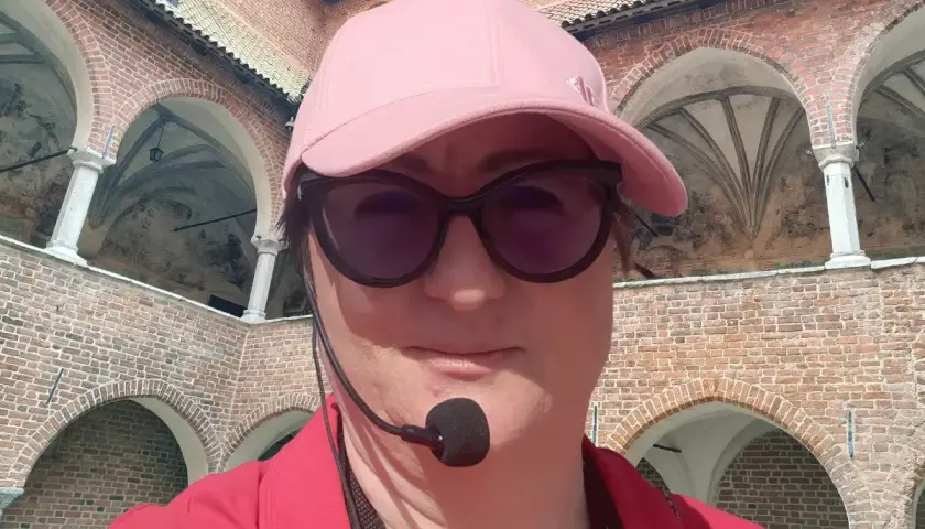 Kobieta w czapce i okularach przeciwsłonecznych zwiedza z przewodnikiem z mikrofonem zamek w Lidzbarku Warmińskim, odkrywając jego atrakcje.
